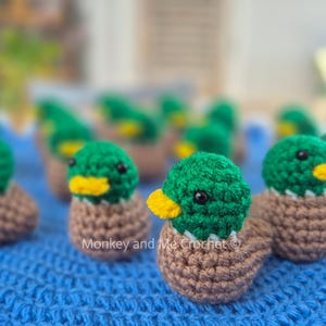 May include: A collection of handmade crochet ducklings. Each duckling features a green head, a yellow beak, and a brown body. The ducklings are arranged on a blue textured surface.