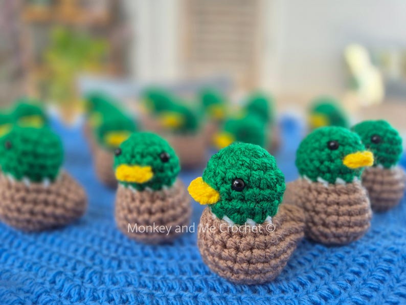 May include: A collection of handmade crochet ducklings. Each duckling features a green head, a yellow beak, and a brown body. The ducklings are arranged on a blue textured surface.