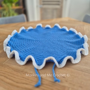 Op de afbeelding: Een rond, blauw gehaakt item met een witte, golvende rand. Het heeft twee blauwe bandjes en de tekst "Monkey and Me Crochet ©". Het item ligt op een houten oppervlak.