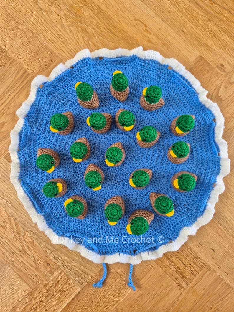 May include: A blue crocheted mat with a white scalloped edge, adorned with numerous crocheted ducklings. Each duckling features a green body, brown wings, and a yellow beak. The text "Monkey and Me Crochet" is visible on the mat.