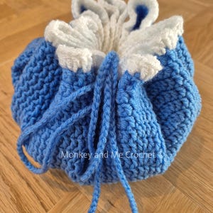 May include: A handmade crochet drawstring bag in shades of blue and white. The bag features a textured stitch pattern and a ruffled, flower-like top. The drawstring is a matching blue yarn. The text "Monkey and Me Crochet" is visible.