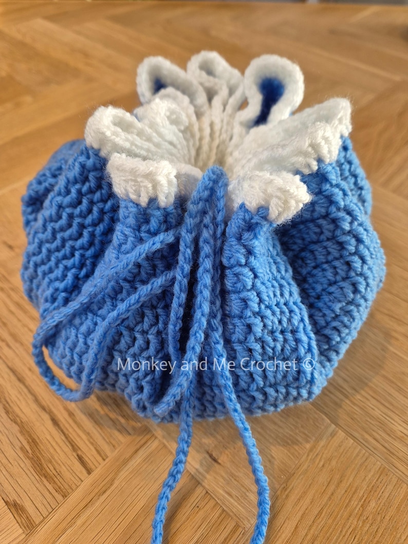 May include: A handmade crochet drawstring bag in shades of blue and white. The bag features a textured stitch pattern and a ruffled, flower-like top. The drawstring is a matching blue yarn. The text "Monkey and Me Crochet" is visible.