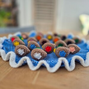 May include: A blue crocheted mat with a white scalloped edge, holding several brown crocheted acorn-shaped objects with colorful details. The acorns have red, yellow, blue, and orange accents. The text "Monkey and Me Crochet" is visible.