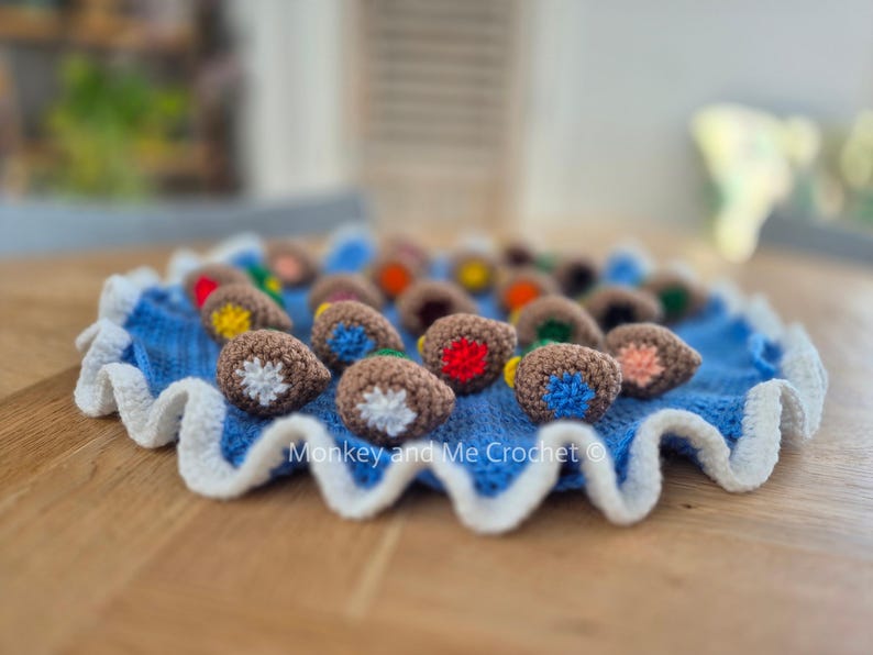 May include: A blue crocheted mat with a white scalloped edge, holding several brown crocheted acorn-shaped objects with colorful details. The acorns have red, yellow, blue, and orange accents. The text "Monkey and Me Crochet" is visible.