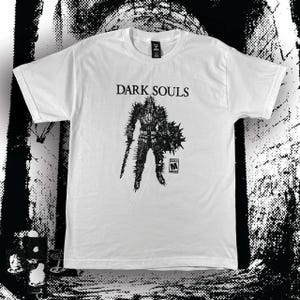 May include: A white short-sleeve t-shirt featuring a black graphic print of a knight in spiked armor holding a sword and shield. The text "DARK SOULS" is printed above the knight, and a small "M" ESRB rating symbol is located below the shield.