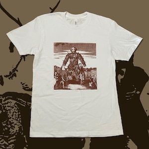 May include: White t-shirt featuring a sepia-toned illustration of a figure with a crown and a group of people. The graphic is centered on the front of the shirt. The shirt has short sleeves and a crew neck.