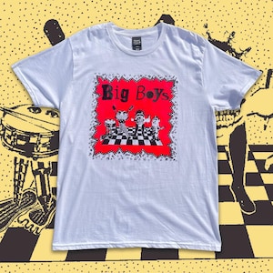 May include: White t-shirt featuring a red square with the words "Big Boys" and a black and white graphic of cartoon figures. The shirt has short sleeves and a crew neck. The background is yellow with black graphic elements.