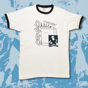 Op de afbeelding: Wit T-shirt met een zwarte boord. Het shirt heeft een zwart-witte afbeelding van de band DEVO met de tekst "Are We Not Men?" en een foto van een man die keyboard speelt.