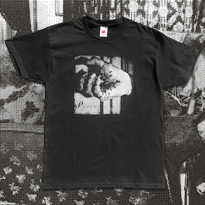 以下が含まれることがあります： 黒いTシャツ。手のグレースケール画像と、その下に「Pixies」の文字がプリントされています。画像は長方形の中の正方形で、手の後ろには縦線があります。クルーネックです。
