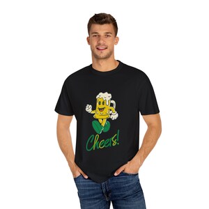 Könnte beinhalten: Schwarzes T-Shirt mit einer Cartoon-Bierkrug-Figur in Gelb, Weiß und Grün. Die Figur hat ein lächelndes Gesicht und geht. Der Text "Cheers!" steht in grüner Schrift darunter. Das T-Shirt wird von einer Person getragen.