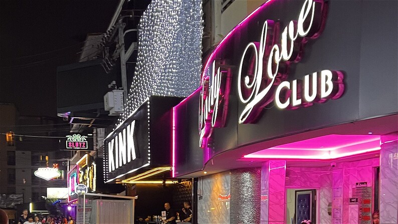 May include: A nighttime street scene with several brightly lit storefronts. The storefront on the right has a pink neon sign that reads "Lady Love Club". The storefront to the left has a sign that reads "Kink".