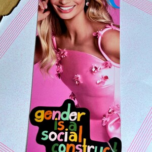 Pode incluir: Uma capa de revista rosa com uma mulher na capa. A mulher está vestindo um vestido rosa com detalhes florais. O texto "gender is a social construct" está na capa em letras coloridas. O título da revista "GOT RO" é visível na parte inferior da capa.