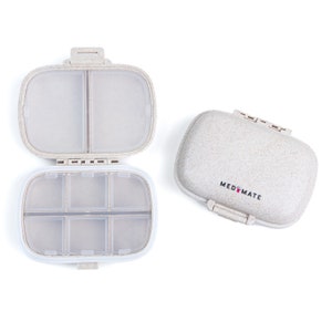 Puede incluir: Un pastillero beige con superficie texturizada, abierto para revelar seis compartimentos. La tapa cerrada muestra el texto "MED MATE" en negro. El pastillero est&aacute; dise&ntilde;ado para almacenar y organizar medicamentos.