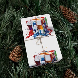 May include: White wrapping paper featuring a colorful, abstract Rubik's Cube design with dripping paint in red, blue, yellow, and green. The paper is tied with twine and set against a backdrop of pine needles and pine cones.