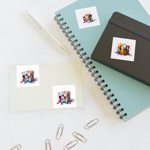 May include: Square stickers featuring a colorful, abstract Rubik's Cube design with paint drips. The cube's faces display various colors, including blue, yellow, red, and white. The stickers are placed on a light blue notebook, a black notebook, and a cream-colored paper.