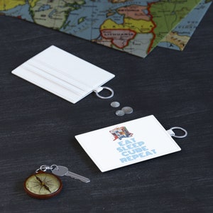 May include: White leather cardholder with a silver keyring, a compass, and a key. The cardholder has the text "EAT SLEEP CUBE REPEAT" printed on it. A world map is in the background.