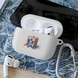 May include: White AirPod case with a colorful graphic of a Rubik's Cube with paint dripping down. The case is open, revealing white earbuds inside. A silver carabiner clip is attached to the case.