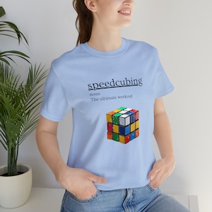 May include: Light blue t-shirt featuring the word "speedcubing" above a colorful Rubik's Cube graphic. The text below the word "speedcubing" reads "noun The ultimate workout". The shirt is made of a soft material.