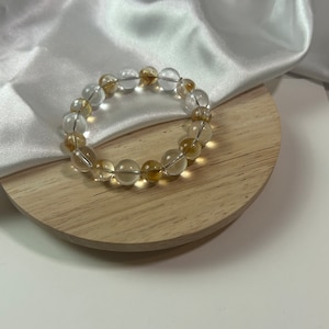 May include: A bracelet made of clear and amber-coloured round beads. The bracelet is on a wooden surface.