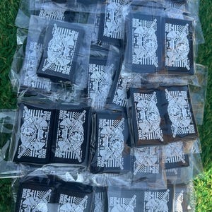 May include: A collection of black rectangular patches, each individually packaged in clear plastic. The patches feature a detailed white graphic design with the text "ROAD DOGZ 2800" and other text. The patches are arranged on a green surface.