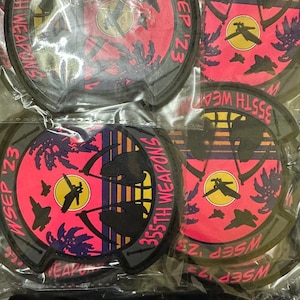 May include: A collection of round patches with a black border. Each patch features a pink and black design with the text "355th Weapons WSEP '23". The design includes silhouettes of aircraft and palm trees.