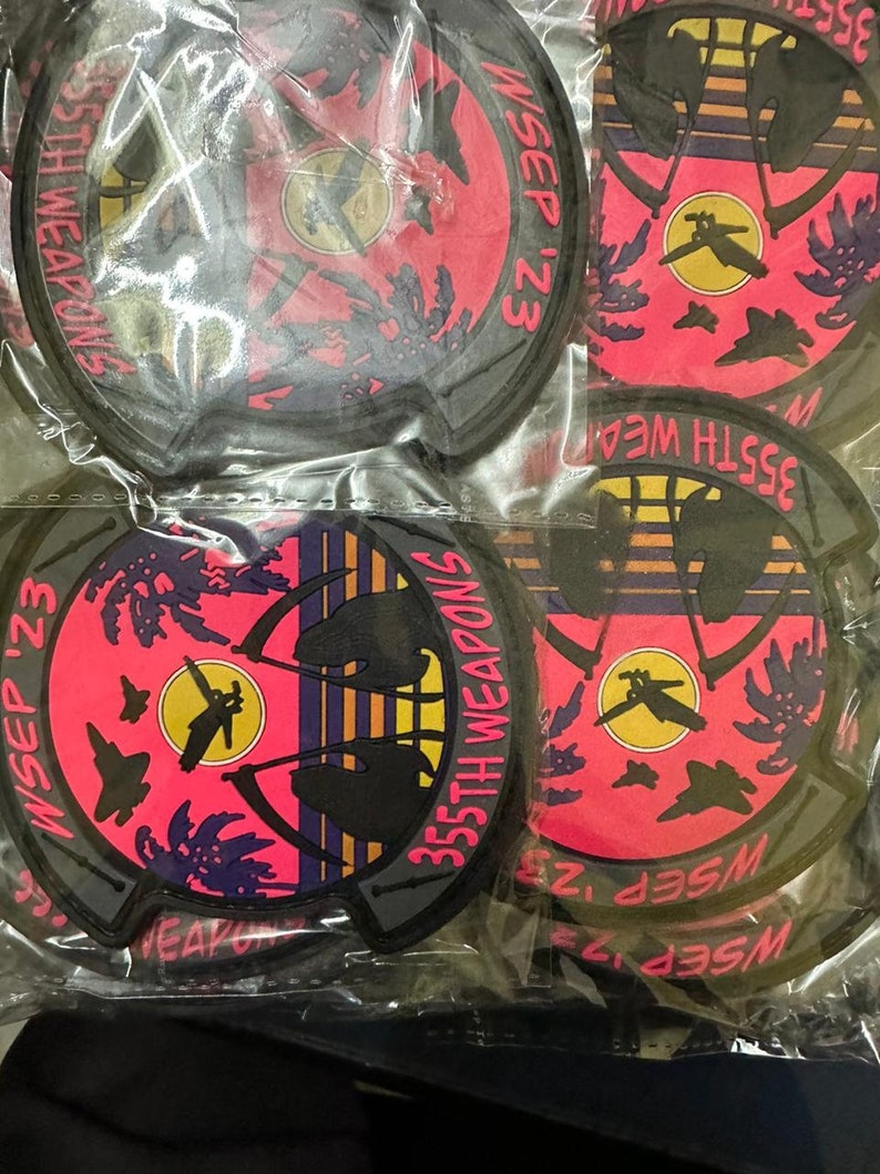 May include: A collection of round patches with a black border. Each patch features a pink and black design with the text "355th Weapons WSEP '23". The design includes silhouettes of aircraft and palm trees.