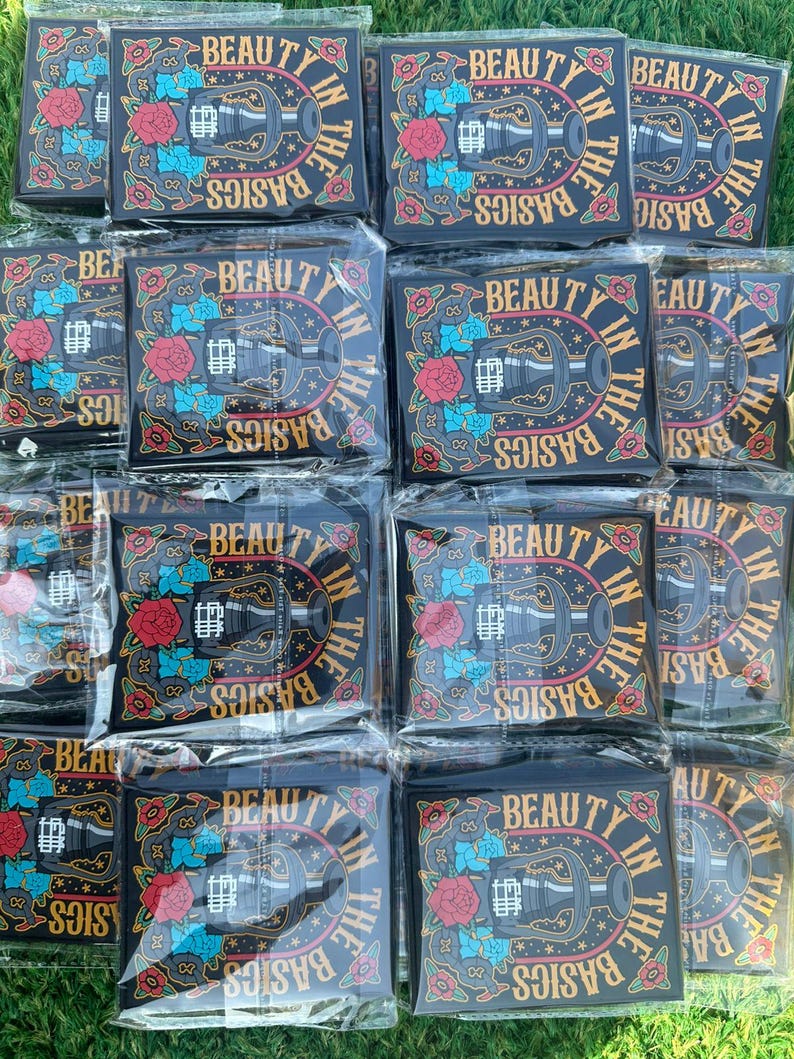 May include: A collection of black patches with the text "BEAUTY IN THE BASICS" in a circular design. The patches feature a central illustration of a bottle, surrounded by red and blue floral accents. The patches are packaged in clear plastic.