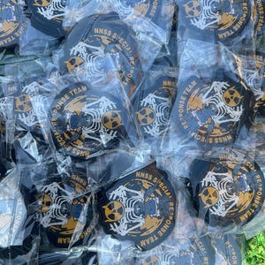 May include: A collection of black and gold patches, each individually packaged in clear plastic. The patches feature a white skeleton design with a radiation symbol and the text "NNSS SPECIAL RESPONSE TEAM".