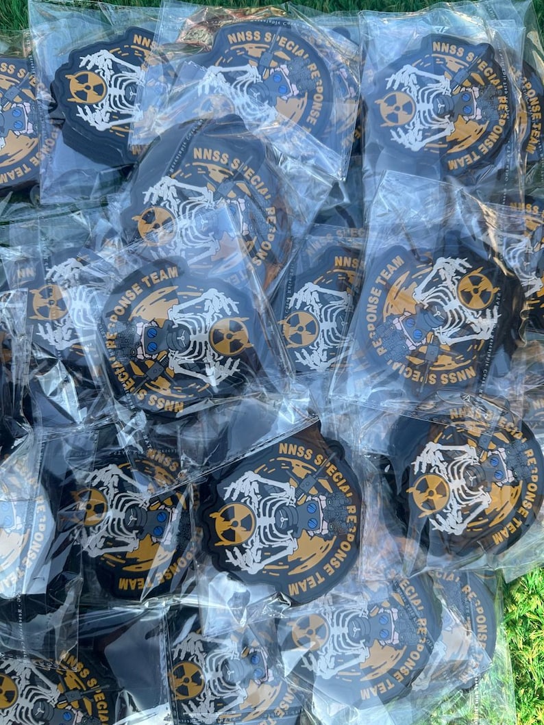 May include: A collection of black and gold patches, each individually packaged in clear plastic. The patches feature a white skeleton design with a radiation symbol and the text "NNSS SPECIAL RESPONSE TEAM".