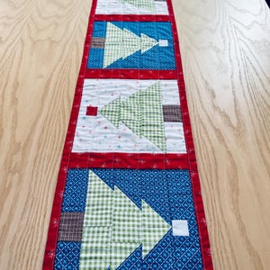 May include: A rectangular Christmas tree table runner. The runner features alternating sections of blue, white, and red fabric. Each section has a patchwork Christmas tree design in green and brown. The runner is approximately 12 inches wide and 48 inches long.