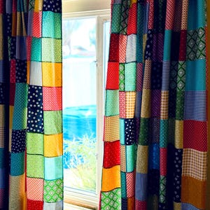 Può includere: Tende patchwork colorate davanti a una finestra. Le tende sono realizzate con vari quadrati di tessuto con motivi rossi, gialli, verdi, blu e bianchi. La finestra rivela una vista di acqua blu e cielo.