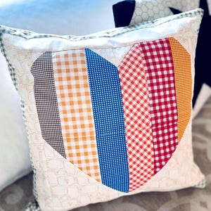 May include: Square decorative pillow with a patchwork heart design. The heart is composed of vertical stripes in various gingham patterns and colors, including brown, orange, blue, and red. The pillow has a cream-colored background with a repeating pattern and a green trim.