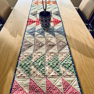 Peut inclure: Une nappe matelassée colorée avec un motif géométrique de triangles rouges, verts, bleus, marrons et oranges. La nappe est sur une table en bois clair, avec une plante en pot au centre. La nappe mesure environ 152 cm de long.