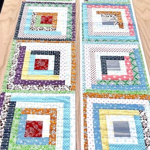 May include: Two colorful, handmade quilted table runners. Each runner features a square-in-square design with a variety of patterned fabrics in shades of red, blue, green, and purple. The runners are approximately 12 inches wide and 48 inches long.