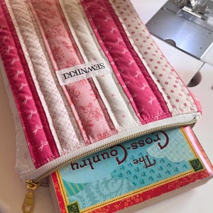 May include: A quilted book sleeve in shades of pink and white, with a gold zipper and the text "SEWNIKKI" on a label. The sleeve holds a book titled "The Cross-Country". The design features vertical stripes and various patterns.