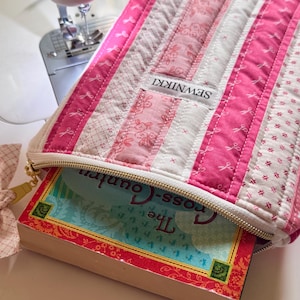 May include: A pink and white quilted book sleeve with a gold zip, holding a book titled "The Cross-Country". The sleeve is made of various patterned fabrics and features the label "SEWNIKKI".