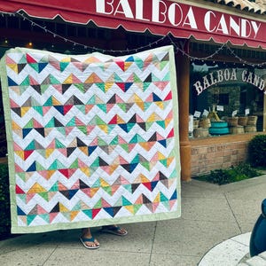 May include: A handmade quilt with a white background and a repeating chevron pattern of colorful triangles. The triangles are in shades of red, blue, yellow, green, and brown. The quilt is bordered with a green and white gingham fabric. The quilt is displayed in front of a candy store.