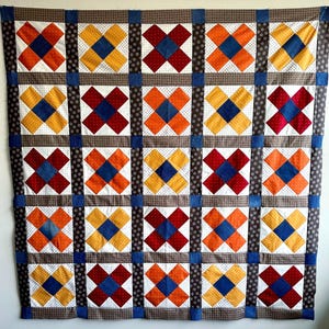 May include: A patchwork quilt featuring a grid of colourful X-shaped blocks. The blocks are made from red, orange, yellow, and blue fabrics, set against a white background. The quilt has a brown border and blue corner accents.