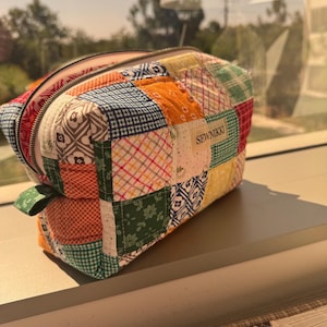 May include: A patchwork quilted cosmetic bag with a zipper closure. The bag features a variety of colorful fabric squares in patterns like gingham, floral, and geometric designs. The label reads "SEWNIKKI".