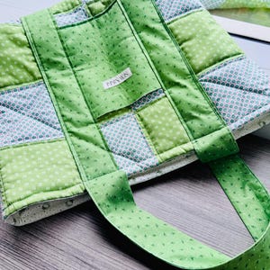 May include: A quilted tote bag in shades of green and white. The bag features a patchwork design with various patterns, including small floral prints and polka dots. The bag has sturdy green straps and a small label that reads "IMMIN WIS".