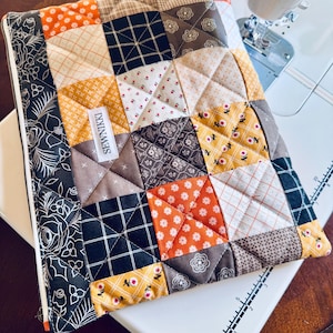 May include: A patchwork quilted laptop sleeve with a variety of fabrics in orange, yellow, brown, and black. The sleeve has a zipper closure and a black floral patterned border. The label reads "SEWNIKKI".