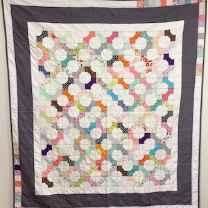 Può includere: Una trapunta fatta a mano con un centro bianco con un motivo colorato di cerchi interconnessi. I cerchi sono riempiti con vari tessuti fantasia nei toni del rosa, arancione, verde e blu. La trapunta è bordata da un tessuto grigio scuro.