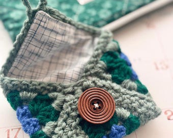 Può includere: Presina all'uncinetto fatta a mano nei toni del verde, blu e grigio, con chiusura a bottone in legno. L'interno è foderato con un tessuto fantasia. Un anello è attaccato per appenderla. La presina è progettata con un motivo a granny square.