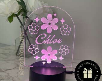 May include: A pink night light with a clear acrylic base and a white arch-shaped panel. The panel has a floral design with the name "Chloe" in the center. The base has a black cord and a black plug.
