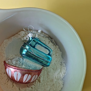 May include: A blue glass Christmas ornament shaped like a vintage kitchen appliance sits in a white mixing bowl filled with white flour.