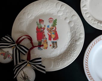 May include: Two white ceramic plates with a Christmas theme. One plate features a raised design of leaves and berries, and a center image of two Santa Clauses carrying gifts. The other plate has a gold border with a repeating pattern.