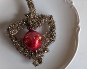 May include: A vintage Christmas ornament shaped like a heart, made of silver tinsel with a red glass ball in the center. The ornament has a wire loop for hanging.