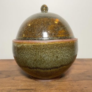 May include: A ceramic dome-shaped lidded bowl with a speckled brown and green glaze. The bowl has a slightly raised rim and a knob on the top of the lid.
