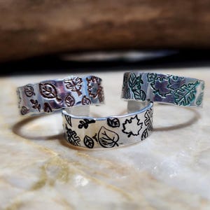 May include: Three silver-toned rings with leaf designs. One ring has a black leaf pattern, another has a brown leaf pattern, and the third has a green leaf pattern. The rings are displayed on a light-colored surface.