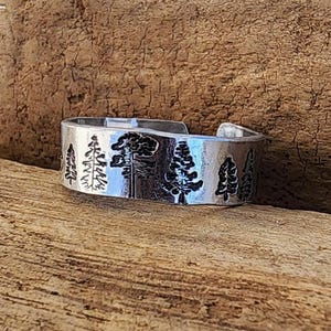 May include: A silver-colored ring with a forest design. The ring features an etched pattern of various trees. The ring is displayed on a wooden surface.